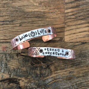 May include: Two copper bracelets with hammered metal surfaces. The top bracelet has the words "have hope" engraved on it. The bottom bracelet has the words "strong & courageous" engraved on it.