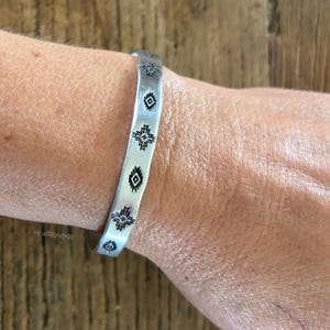 May include: A silver-toned metal cuff bracelet featuring a series of stamped black geometric patterns along its length. The bracelet has a smooth, flat surface with repeating diamond and cross-shaped motifs etched into the metal, creating a distinct visual contrast against the silver finish.