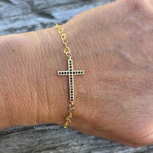 May include: Gold-toned bracelet featuring a cross pendant adorned with black gemstones. The bracelet's chain incorporates heart-shaped links. The image shows the bracelet worn on a wrist.