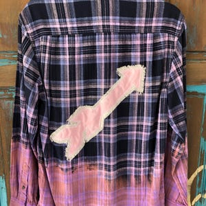 May include: A long-sleeve button-down flannel shirt featuring a black and light purple plaid pattern. The bottom portion of the shirt is bleached to a faded pinkish-orange hue. A large, light pink fabric arrow patch with frayed edges is sewn on the back.