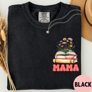 May include: A black crewneck t-shirt featuring a graphic of three stacked books with a bouquet of colorful wildflowers emerging from the top book. Below the books, the word MAMA is printed in a bold, pink, retro-style font. A silver star necklace rests on the collar.