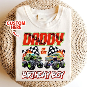 May include: Cream-colored t-shirt featuring the text "DADDY OF THE BIRTHDAY BOY" in red and green. Two monster trucks with checkered flags are also displayed. The words "CUSTOM HERE" are in the upper left corner.