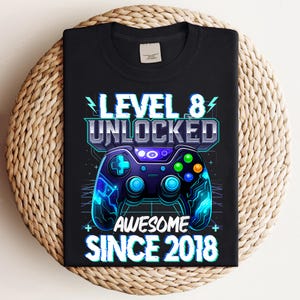 May include: Black t-shirt with a neon blue and purple video game controller graphic. The text "LEVEL 8 UNLOCKED AWESOME SINCE 2018" is displayed in a futuristic font. The design incorporates lightning bolt graphics.