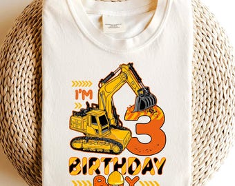 May include: Cream-coloured t-shirt with a yellow excavator graphic and the number 3 in orange. Text reads "I'm" and "Birthday Boy" in yellow and orange, with a construction hat icon. The shirt is on a woven background.