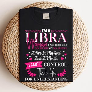 May include: Black t-shirt with the text "I'M A LIBRA Woman I Was Born With MY HEART ON MY SLEEVE A Fire In My Soul And A Mouth I CAN'T CONTROL Thank You FOR UNDERSTANDING" in white and pink. The shirt is displayed on a woven surface.