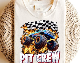 May include: Cream-colored t-shirt featuring a blue monster truck design with orange and black tires, flames, and a checkered flag. The text "PIT CREW" is printed in white with a red outline. The shirt is folded on a woven surface.