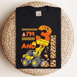以下が含まれることがあります： 建設をテーマにした黒いTシャツ。デザインには、黄色のフロントエンドローダー、オレンジ色の数字3、オレンジと黄色のフレーズ「I'm and Digging It！」が含まれています。Tシャツは織られた表面にあります。
