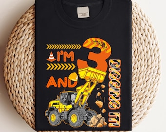 May include: Black t-shirt with a construction theme. The design features a yellow front-end loader, the number 3 in orange, and the phrases "I'm and Digging It!" in orange and yellow. The shirt is on a woven surface.