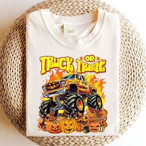 May include: Cream-colored t-shirt featuring a Halloween-themed graphic. The design includes a monster truck with flames, the words "Truck or Treat," and jack-o'-lanterns. The shirt is made of a soft material.