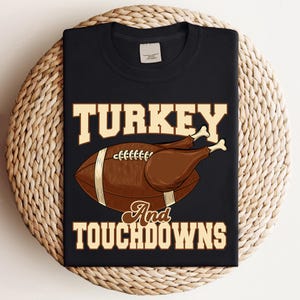 Pode incluir: T-shirt preto com um gráfico que apresenta uma bola de futebol americano e uma coxa de peru. O texto diz "TURKEY And TOUCHDOWNS" em uma fonte serif. A bola é marrom com listras brancas, e a coxa de peru é marrom com ossos brancos.