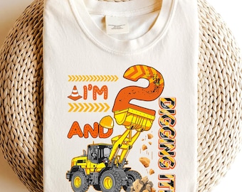 May include: Cream-coloured t-shirt with a construction theme. The design features a yellow and black front-end loader, the number "2", and the phrases "I'm and Digging It!" in orange and black. Suitable for a child's birthday.