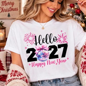 Pode incluir: Uma t-shirt branca com o texto preto Hello 2027, onde uma bola de discoteca rosa substitui o zero. O design inclui gráficos de fogo de artifício rosa e o texto rosa Happy New Year, acentuado com um laço rosa na bola.