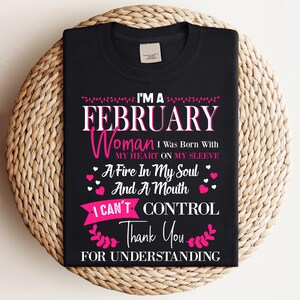 Könnte beinhalten: Schwarzes T-Shirt mit dem Text "I'M A FEBRUARY Woman I Was Born With MY HEART ON MY SLEEVE A Fire In My Soul And A Mouth I CAN'T CONTROL Thank You FOR UNDERSTANDING" in weißer und rosa Schrift. Das T-Shirt wird auf einer gewebten Oberfläche präsentiert.