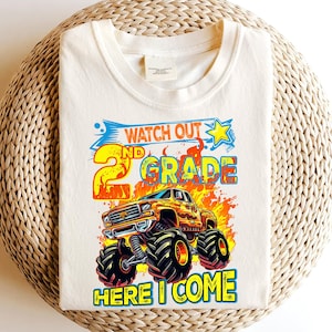 Könnte beinhalten: Cremefarbenes T-Shirt mit einem farbenfrohen Grafikdesign. Das Design zeigt den Text "WATCH OUT 2ND GRADE HERE I COME" und eine Monster-Truck-Illustration. Text und Truck sind in Orange-, Gelb- und Blautönen gehalten.