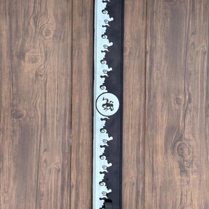 Op de afbeelding: Zwarte en witte gitaarband met een herhalend wolkenachtig patroon langs de randen. Een cirkelvormig embleem met een gestileerd ontwerp is gecentreerd op de band. De band heeft een zwarte verstelgesp.