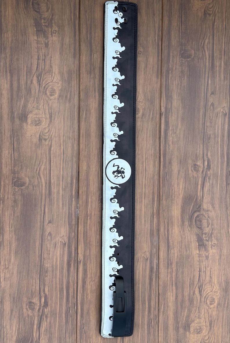 Op de afbeelding: Zwarte en witte gitaarband met een herhalend wolkenachtig patroon langs de randen. Een cirkelvormig embleem met een gestileerd ontwerp is gecentreerd op de band. De band heeft een zwarte verstelgesp.