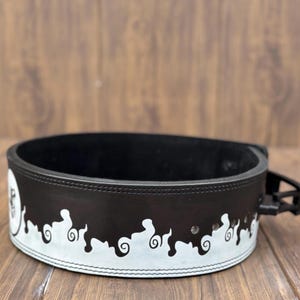 Op de afbeelding: Een zwarte gewichthefband met een wit decoratief ontwerp langs de onderrand. De band is gemaakt van dik leer en heeft een gespsluiting. Het ontwerp heeft een reeks gestileerde, wervelende vormen.