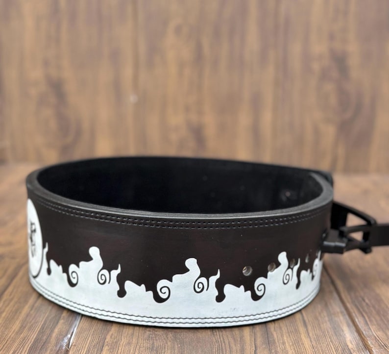 Op de afbeelding: Een zwarte gewichthefband met een wit decoratief ontwerp langs de onderrand. De band is gemaakt van dik leer en heeft een gespsluiting. Het ontwerp heeft een reeks gestileerde, wervelende vormen.