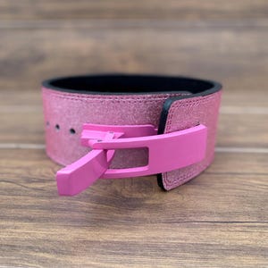 May include: A pink weightlifting belt with a glittery finish and a black interior. The belt features a bright pink buckle and is designed for support during heavy lifting exercises. The belt is shown on a wooden surface.