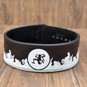 Op de afbeelding: Een zwarte gewichthefband met een wit decoratief ontwerp. De band heeft een wit cirkelvormig embleem met een gestileerde figuur. De band is gemaakt van leer en heeft een gespsluiting.