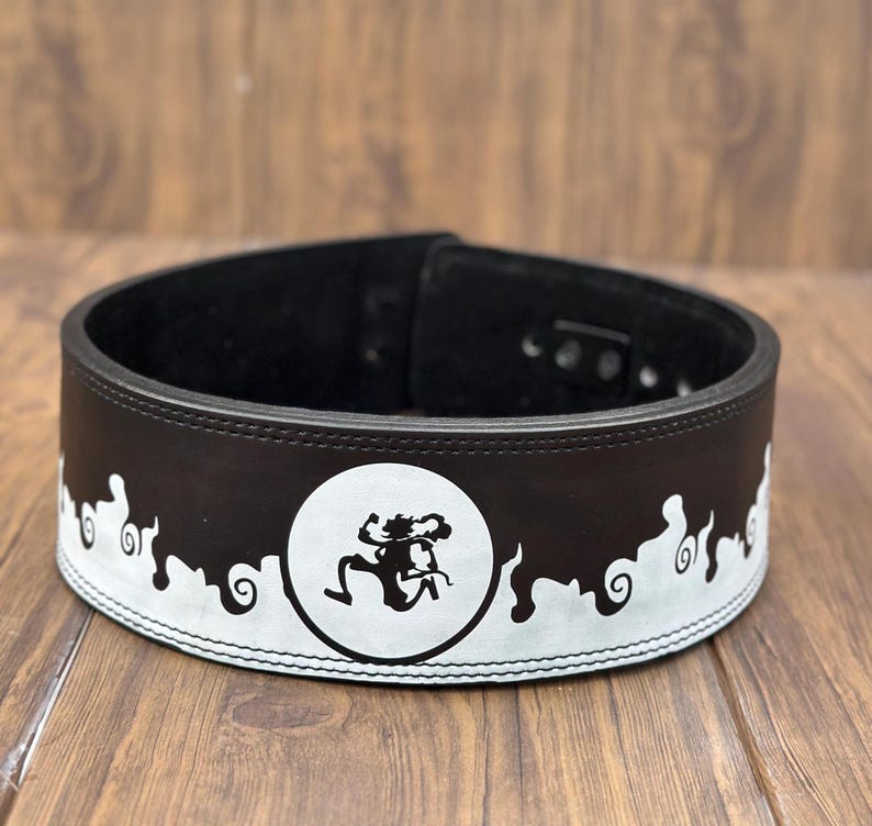 Op de afbeelding: Een zwarte gewichthefband met een wit decoratief ontwerp. De band heeft een wit cirkelvormig embleem met een gestileerde figuur. De band is gemaakt van leer en heeft een gespsluiting.