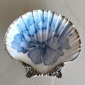 May include: A decorative scallop shell dish featuring a blue and white floral illustration on the interior surface. The shell has a gold-toned rim and is supported by a metallic base with ornate, filigree-style feet at the bottom edge.