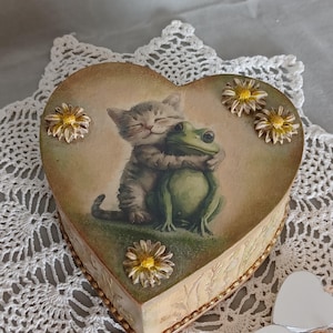 Puede incluir: Una caja en forma de corazón con una ilustración pintada de un gatito atigrado gris abrazando a una rana verde. La tapa está decorada con tres pequeñas flores de margarita blancas y amarillas en relieve. Los lados tienen un acabado texturizado.