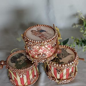Op de afbeelding: Drie cilindervormige trommelornamenten met vintage illustraties aan de bovenkant. Eén toont twee kinderen, een andere een hond en de derde een kat. De zijkanten zijn versierd met rood-witte strepen of patronen, gouden koorden en kralen.