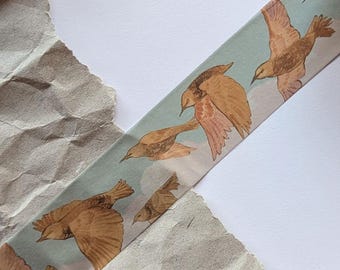 May include: Washi tape with a repeating pattern of brown birds in flight against a light blue sky. The tape is partially rolled, showing the design. The background includes crumpled brown paper and white space.