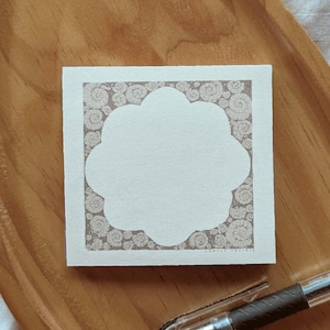 May include: A square, off-white notepad with a decorative border of spiral shell designs. The center of the notepad has a unique, scalloped shape. A black pen rests near the notepad on a wooden tray. The text "LONELY PRAIRIE" is visible.