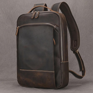May include: A dark brown leather backpack with multiple zippered compartments. The backpack has a rectangular shape with padded shoulder straps and a top handle. The stitching is a lighter brown color, and the zippers are silver.