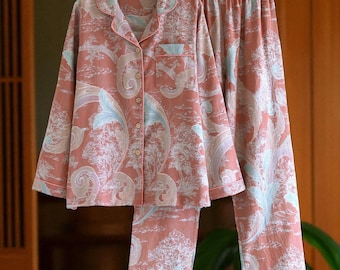 May include: A two-piece pajama set in a coral color with a detailed, repeating pattern of trees and foliage in white and light blue. The long-sleeved top has a button-down front and a chest pocket. The pants have a drawstring waist.