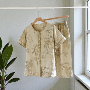 May include: A beige pajama set with a bamboo print, hanging on a wooden hanger. The short-sleeved top has a button-down front and a chest pocket. The matching pants are visible. The set is made of a lightweight fabric.