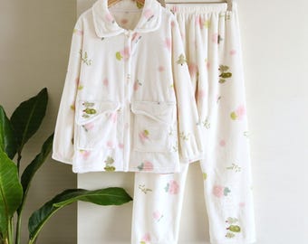 May include: A white pajama set with a floral pattern. The set includes a long-sleeved button-up top with two pockets and pants. The floral design features pink and green accents. The pajamas are hanging on a wooden hanger.