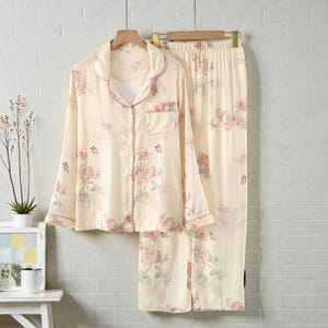 May include: Cream-colored pajama set with a floral pattern in pink and green. The long-sleeved button-down shirt and matching pants feature a delicate print of flowers and butterflies. The set is trimmed with a pink border.