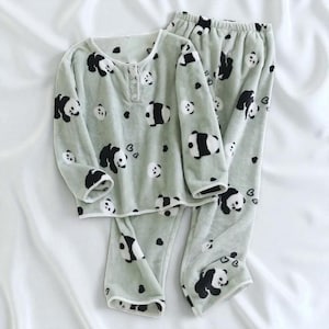 May include: A light green pajama set featuring a repeating panda bear pattern in black and white. The long-sleeved top has a button-up neckline, and the pants have an elastic waistband. The soft, plush fabric is ideal for lounging.