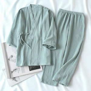 Può includere: Un completo pigiama verde chiaro, composto da un top avvolgente a maniche lunghe con chiusura a fiocco e pantaloni abbinati. Il completo è realizzato in tessuto strutturato ed è esposto su una superficie bianca. Un libro con illustrazioni è parzialmente visibile.