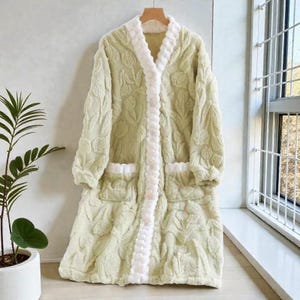 May include: A light green, plush bathrobe with a floral pattern and white trim. The robe has a shawl collar, two front pockets, and a matching belt. It is hanging on a wooden hanger.