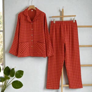 Puede incluir: Un conjunto de pijama de dos piezas con un estampado de cuadros rojos y naranjas. La parte superior de manga larga tiene cuello, cierre de botones y dos bolsillos delanteros. Los pantalones a juego tienen cintura elástica. El conjunto está colgado en una percha de madera.