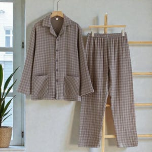 May include: A two-piece pajama set in a neutral brown plaid pattern. The top has a collared neckline, button-down front, and two front pockets. The pants have an elastic waistband. The set is hanging on a wooden hanger.