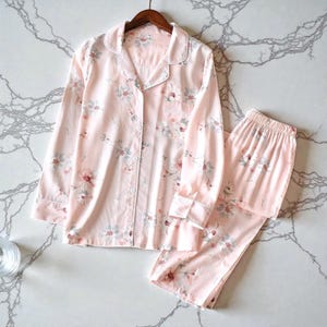 May include: A light pink pajama set with a floral pattern. The set includes a button-down shirt with a chest pocket and long pants. The floral design features pink, gray, and white flowers. The pajamas are made of a soft, lightweight fabric.