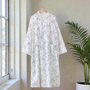 May include: White kimono-style robe with a floral pattern in black. The robe has a V-neck, long sleeves, and pockets. It is hanging on a wooden hanger against a neutral wall, with a potted plant in the background.