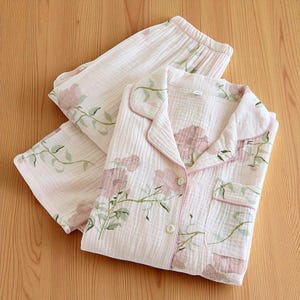 Può includere: Un completo pigiama rosa chiaro con un motivo floreale. Il set include una camicia abbottonata con taschino e pantaloni. Il design floreale presenta fiori rosa e foglie verdi. I bordi della camicia sono rifiniti con un bordo rosa.