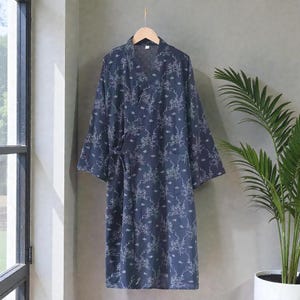 Può includere: Una vestaglia blu scuro in stile kimono con un motivo floreale bianco, appesa a un'appendiabiti in legno. La vestaglia ha maniche lunghe e un design avvolgente con chiusura a legaccio. Lo sfondo è un muro grigio neutro con una finestra e una pianta in vaso.