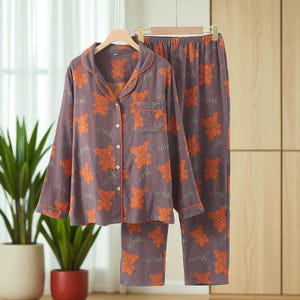 Peut inclure: Un ensemble de pyjama deux pièces violet, orné d'un motif répété d'ours en peluche orange et du mot "wink". L'ensemble comprend une chemise boutonnée avec une poche poitrine et un pantalon assorti. Le pyjama est suspendu à un cintre en bois.
