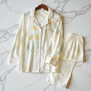 May include: Cream-colored pajama set with a floral pattern. The long-sleeved top has a button-down front, a chest pocket, and white trim. The matching pants have an elastic waistband. The floral design features yellow flowers and green leaves.