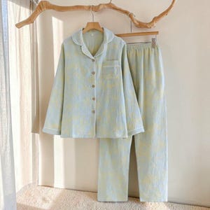 Puede incluir: Un conjunto de pijama azul claro con un estampado floral amarillo. El conjunto incluye una camisa con botones con cuello de solapa y un bolsillo en el pecho, y pantalones a juego. La camisa y los pantalones tienen ribetes blancos. El pijama está colgado en una percha de madera.