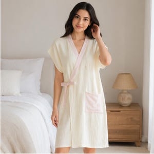 Pode incluir: Um roupão de cor creme de manga curta com design cruzado, debrum rosa claro ao longo do decote e um laço de fecho a condizer na cintura. A peça inclui um bolso de remendo de cor rosa claro na parte da frente.