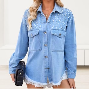 Puede incluir: Chaqueta de camisa vaquera azul claro con adornos de perlas en los hombros. La chaqueta tiene dos bolsillos en el pecho, cierres de botón y un dobladillo deshilachado. Se sostiene un clutch negro.