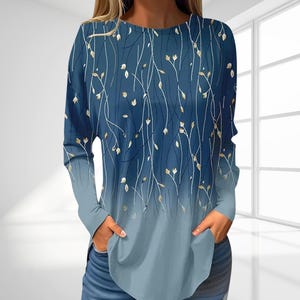Könnte beinhalten: Langärmliges Oberteil mit blauem und hellblauem Farbverlauf. Das Oberteil hat ein Blumenmuster aus weißen Stielen und goldfarbenen Blättern. Das Model trägt eine blaue Jeans und hat die Hände in den Taschen des Oberteils.